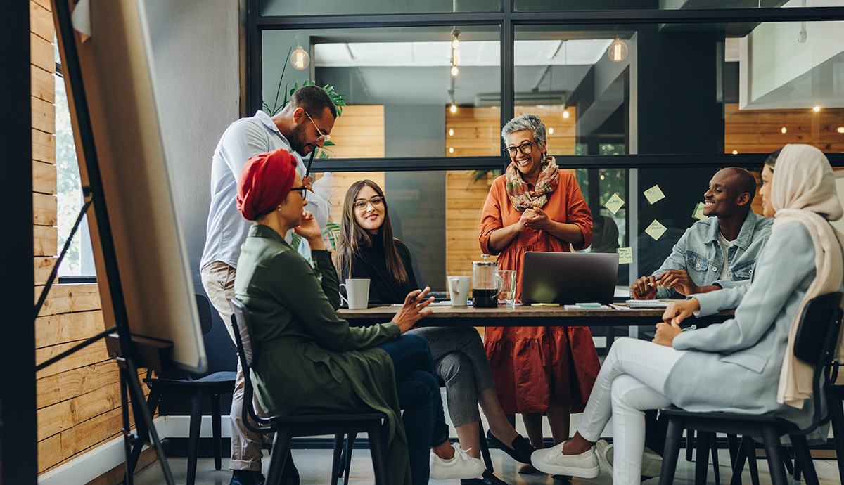 Kulturwandel ermöglichen // Multicultural team of businesspeople smiling during a meeting at the office, while using a laptop and drinking coffee
